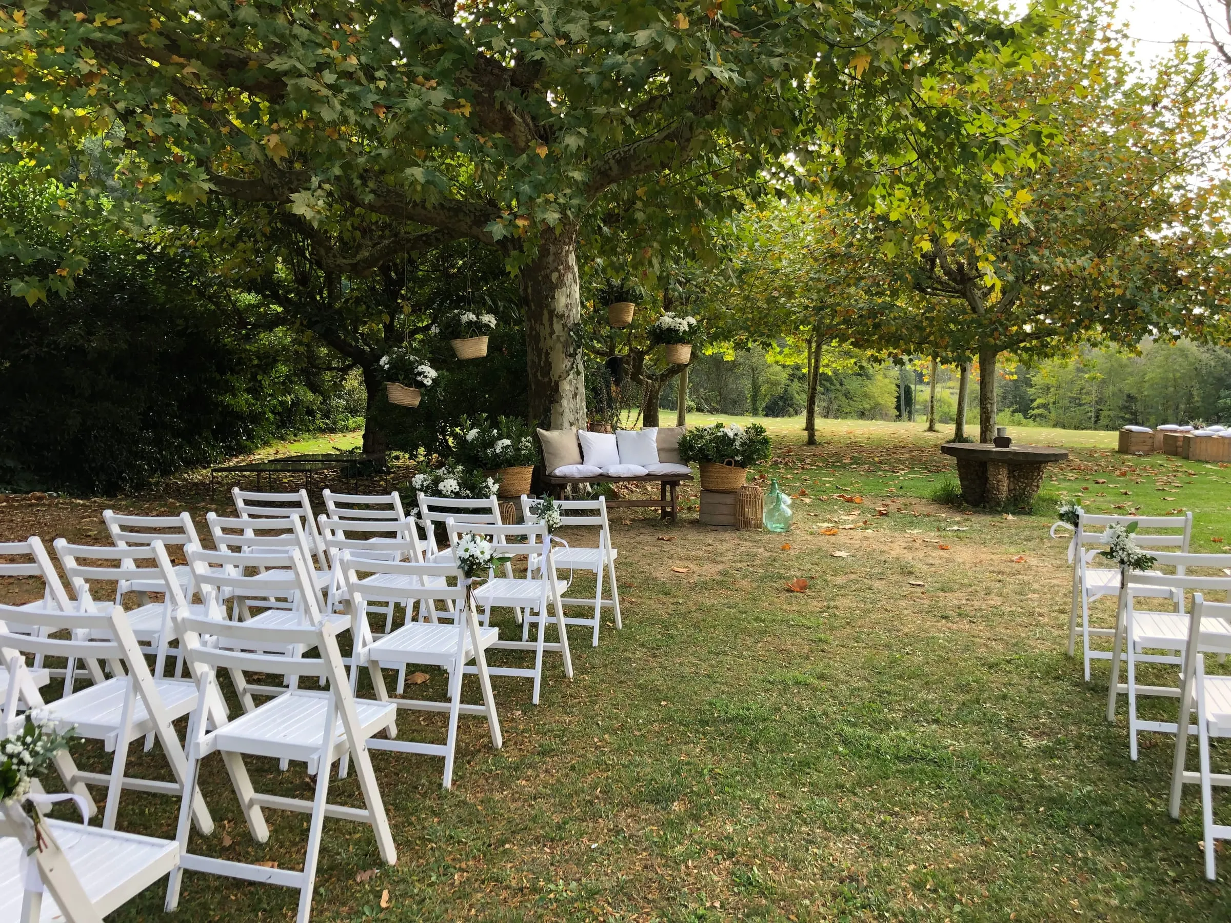 Rows of white folding chairs on the lawn facing a bench beneath the trees