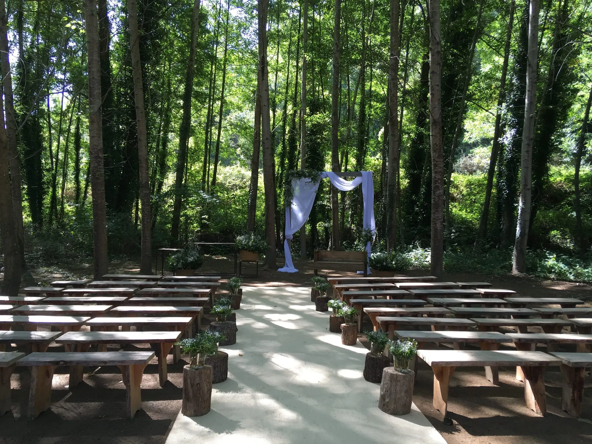 Ceremony in the woods by the stream, with wooden benches and a white fabric arch