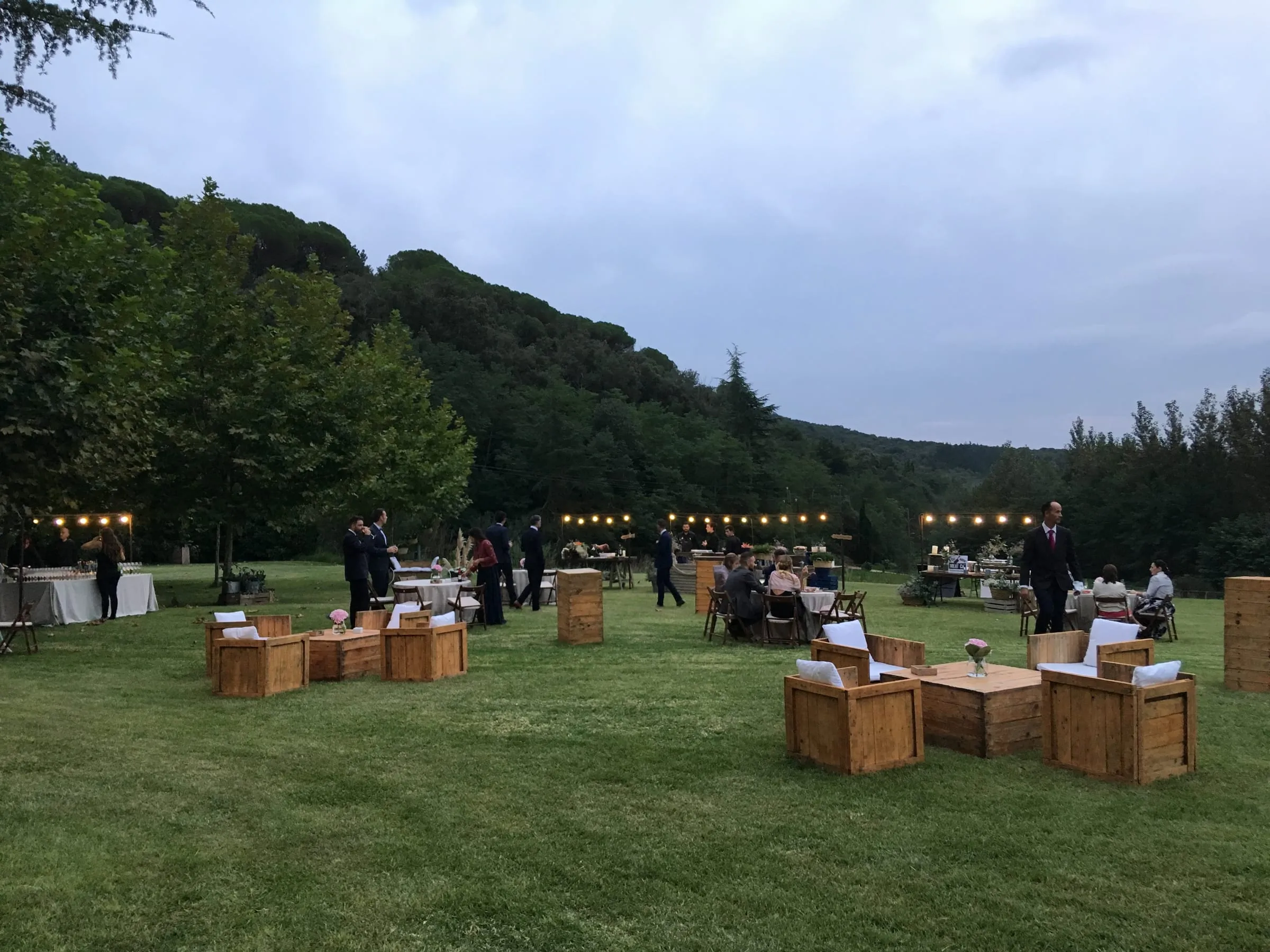 Cocktail hour on the lawn at dusk, with low wooden furniture and strings of lights