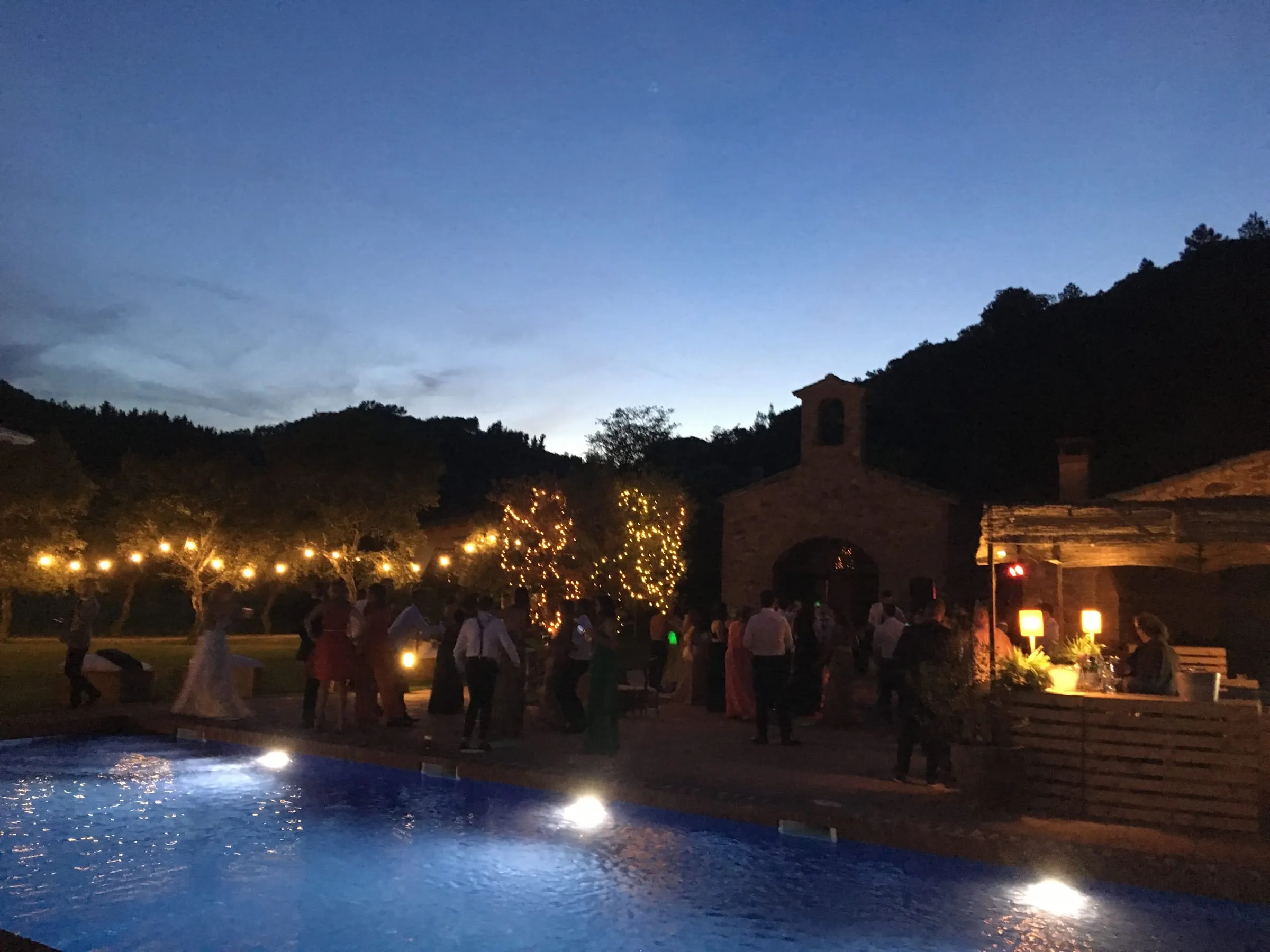 Guests celebrating at nightfall by the lit pool, with the farmhouse and strings of lights behind