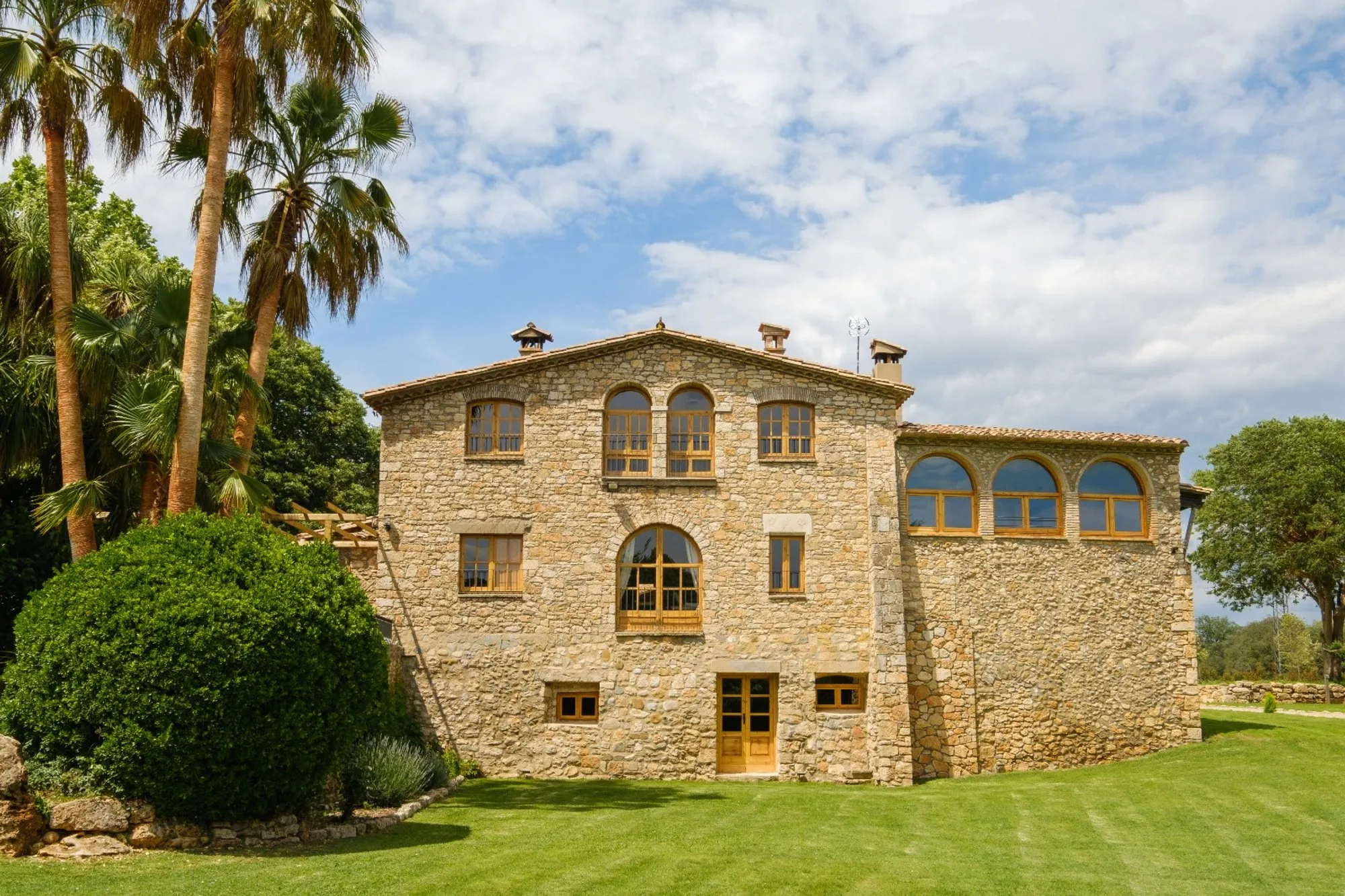 Façana de pedra de la masia Mas Cufí amb finestres en arc, palmeres i gespa al davant
