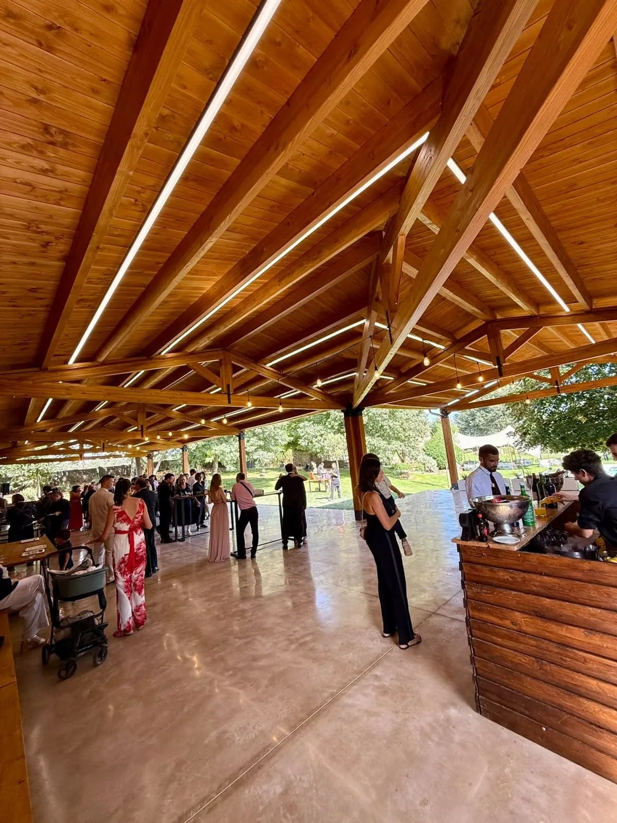 Porche cubierto de madera con una barra y luces en el techo, lleno de invitados