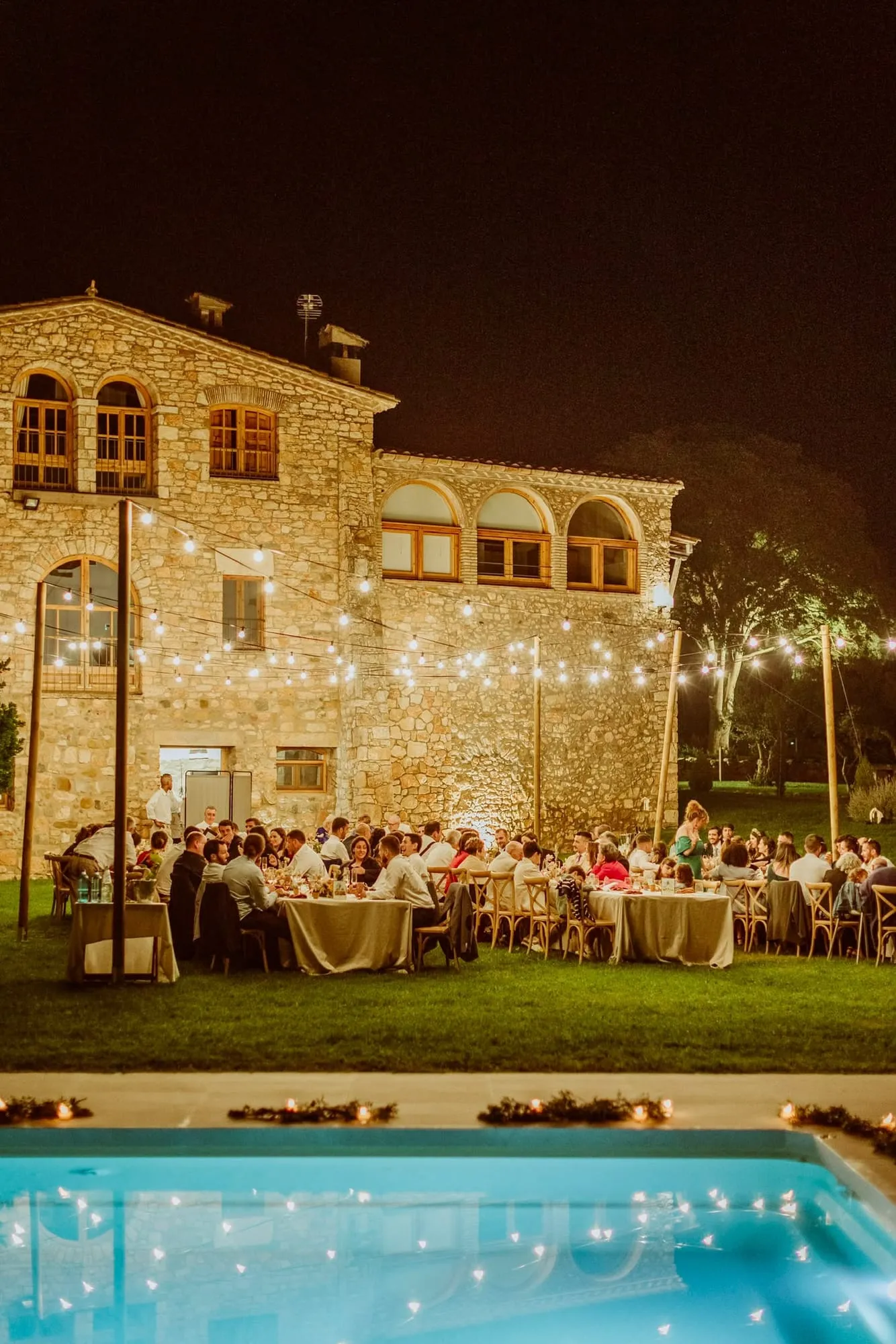 Evening dinner in front of the lit-up farmhouse, with strings of bulbs above the tables