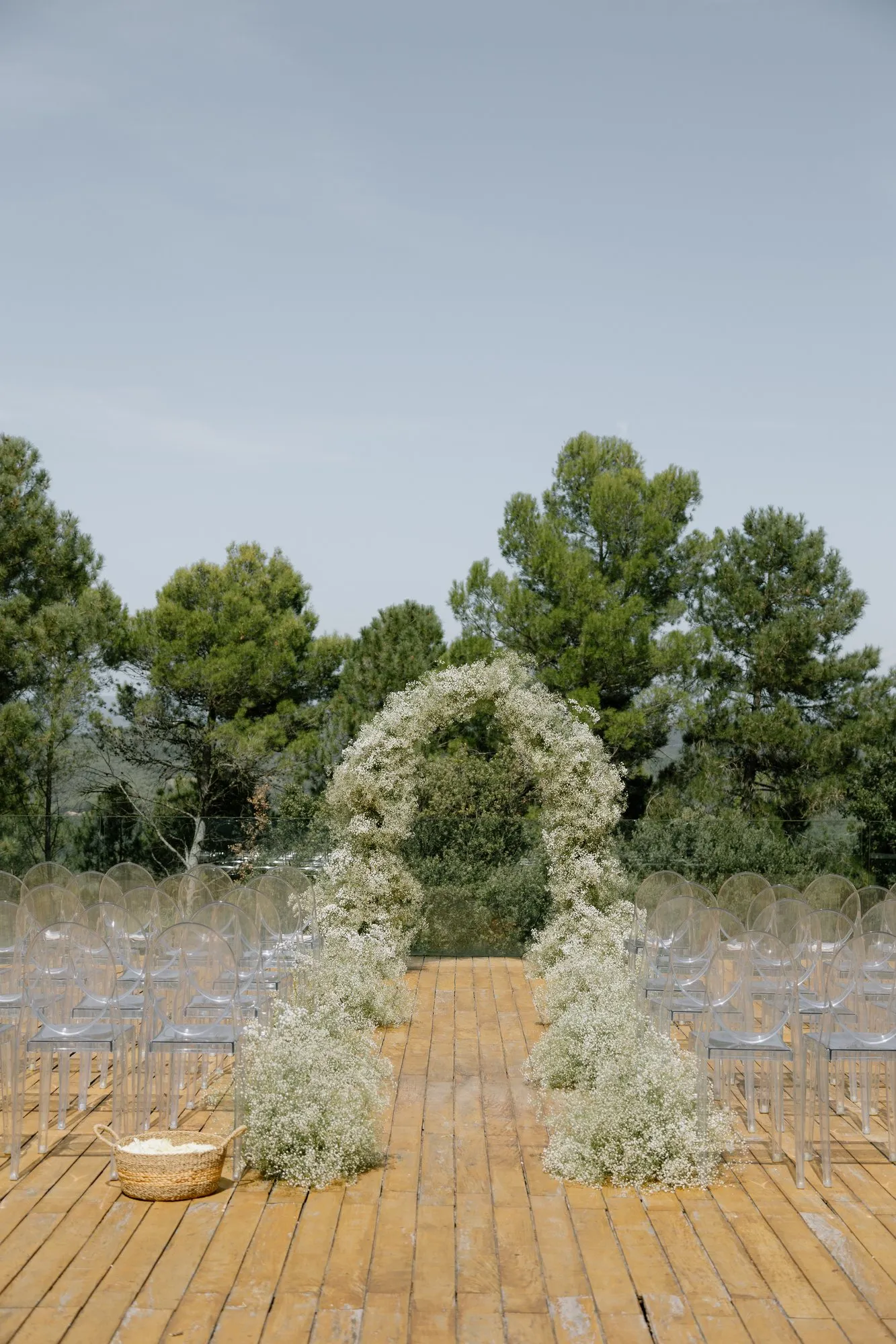 Arch of white flowers on a wooden platform for the ceremony, with chairs and pine trees around it