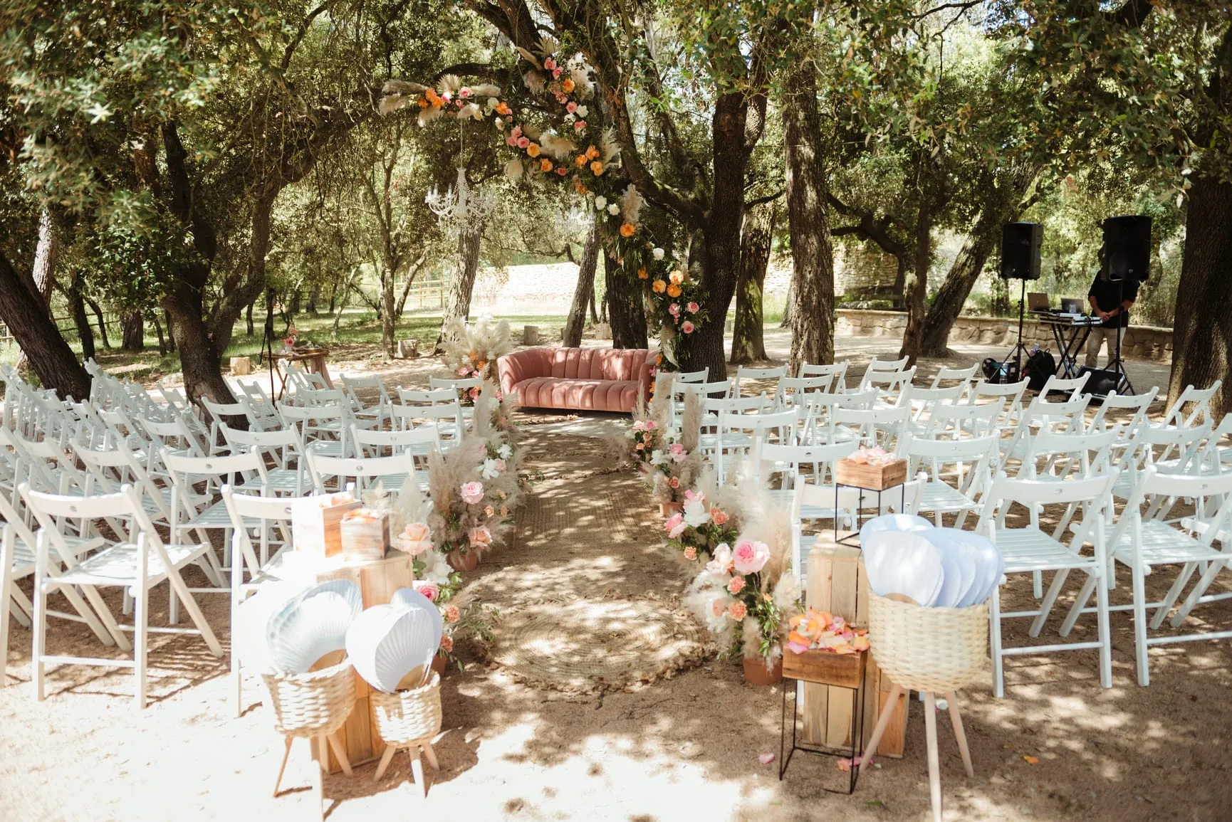 White folding chairs under the holm oaks, with an aisle leading to a pink sofa and a flower arch for the ceremony