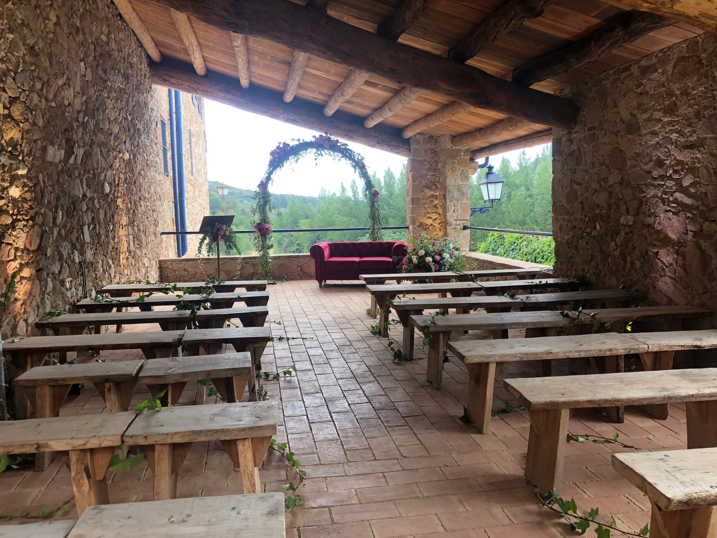 Porxo cobert amb bancs de fusta davant d'un arc de flors i vistes al bosc
