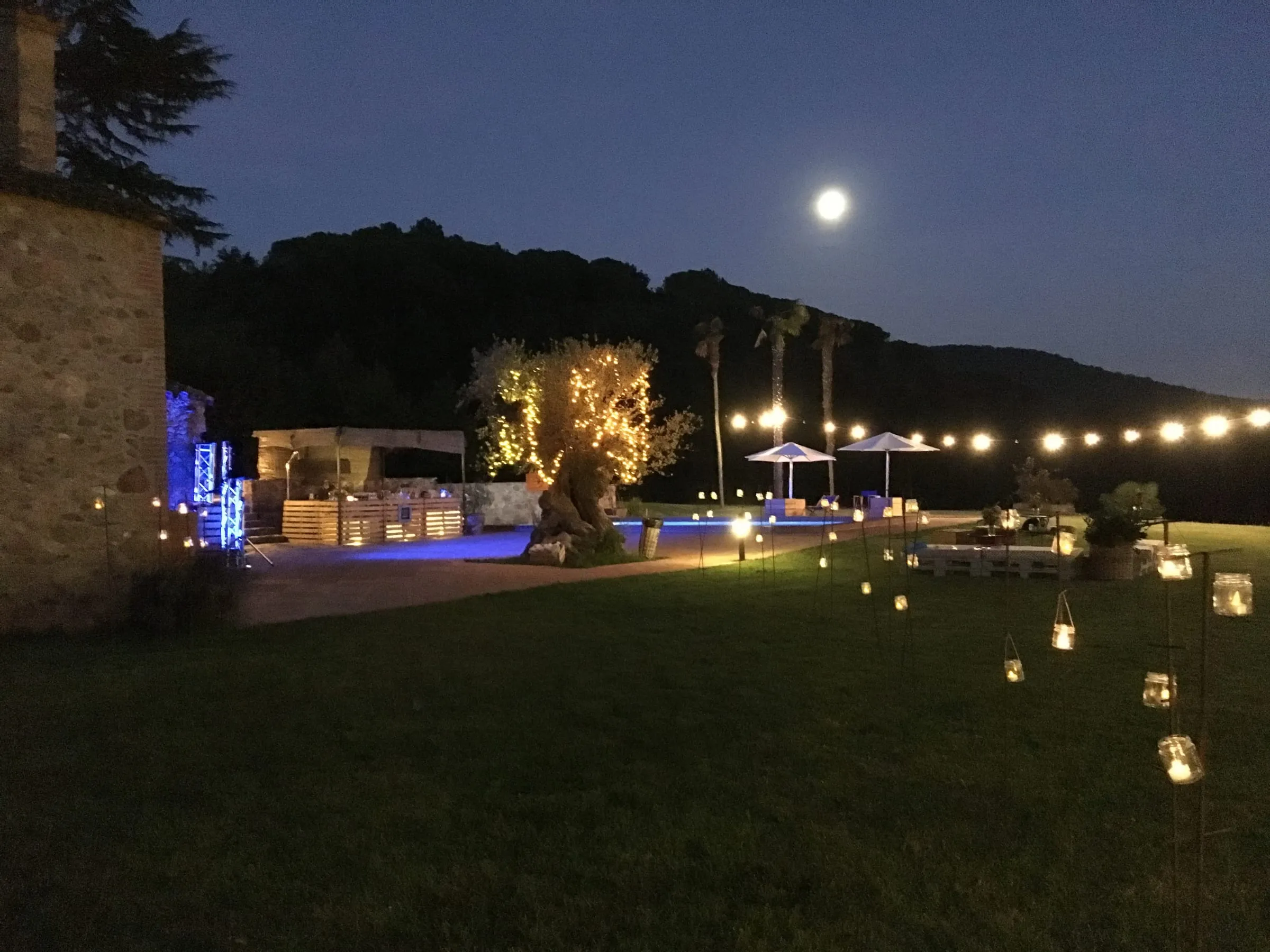 Garden at night with the pool and trees lit up and the moon above the hills